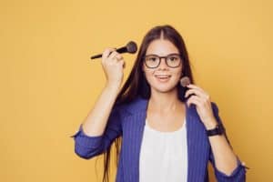woman in glasses holding makeup brushes next to face